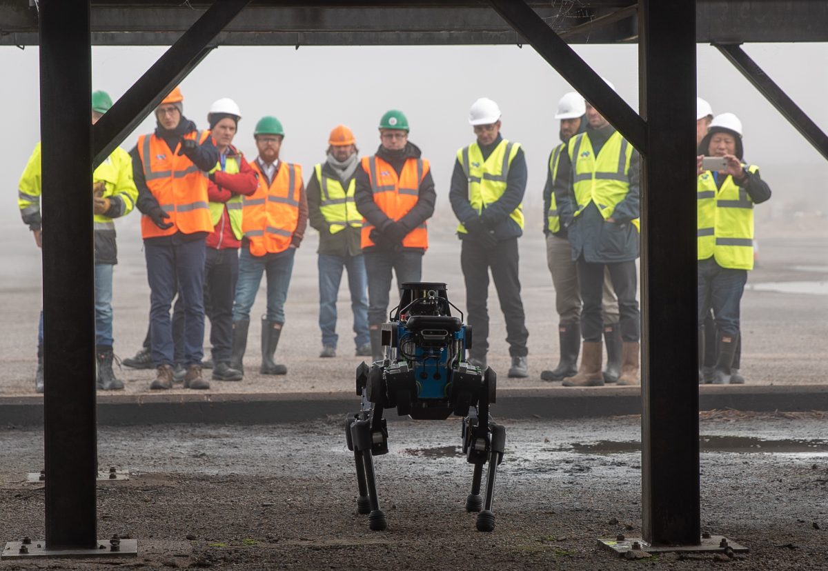 ANYmal B standing in front at industrial site, group in hivis jackets behind. 