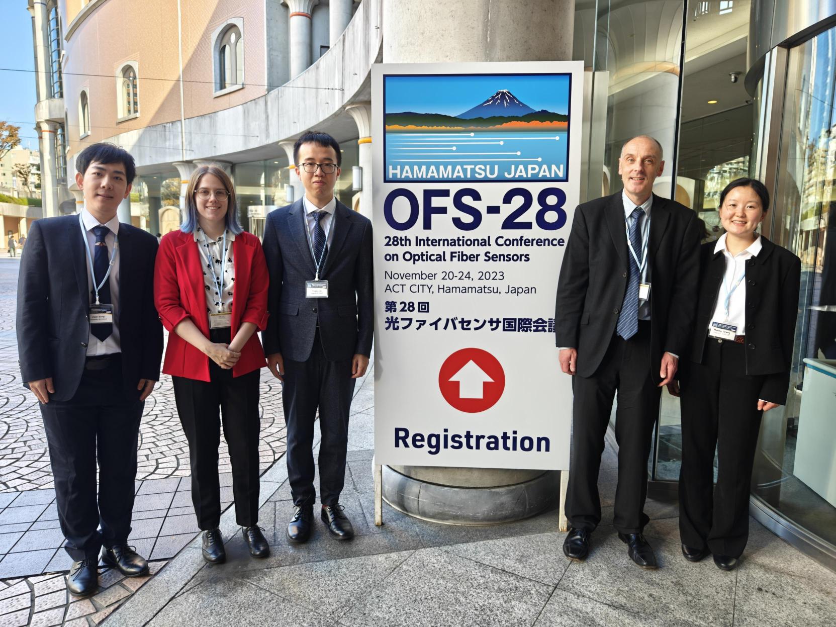 Oxford Team: five people are stood by a conference sign outside of a building. Three people stand on the left of the sign and two on the right, all are wearing business clothes suitable for a conference. On the left, we have an East Asian man, a white woman, and another East Asian man. Then on the right, we have a white man and an Asian woman.