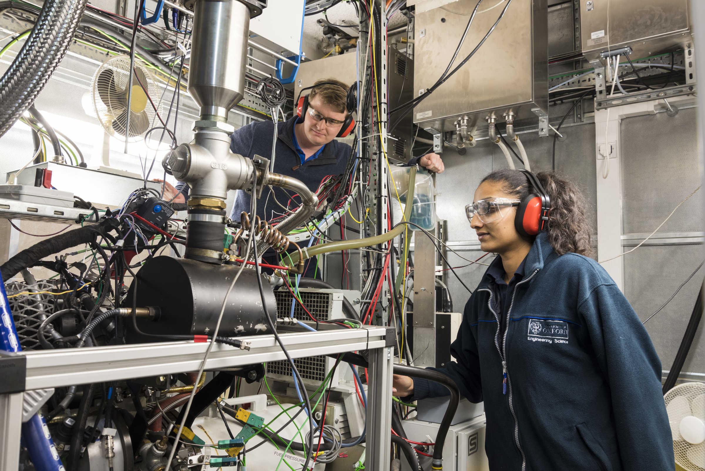 Two students with ear protectors and protective eye glasses look at a big complicated machine