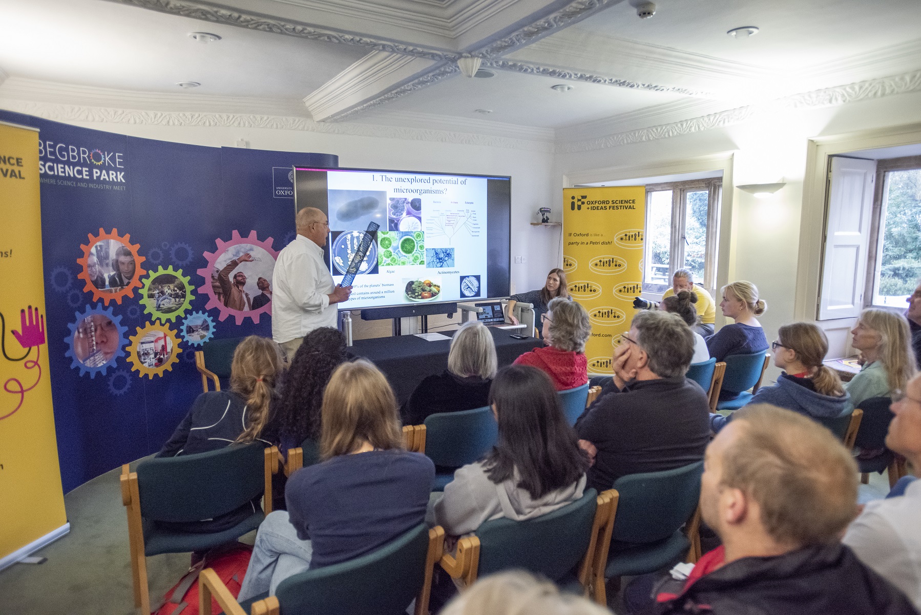 A low resolution image of Professor Ian Thompson stood in front of roller banners and a screen with a PowerPoint presentation on it, presenting to a small room of people.