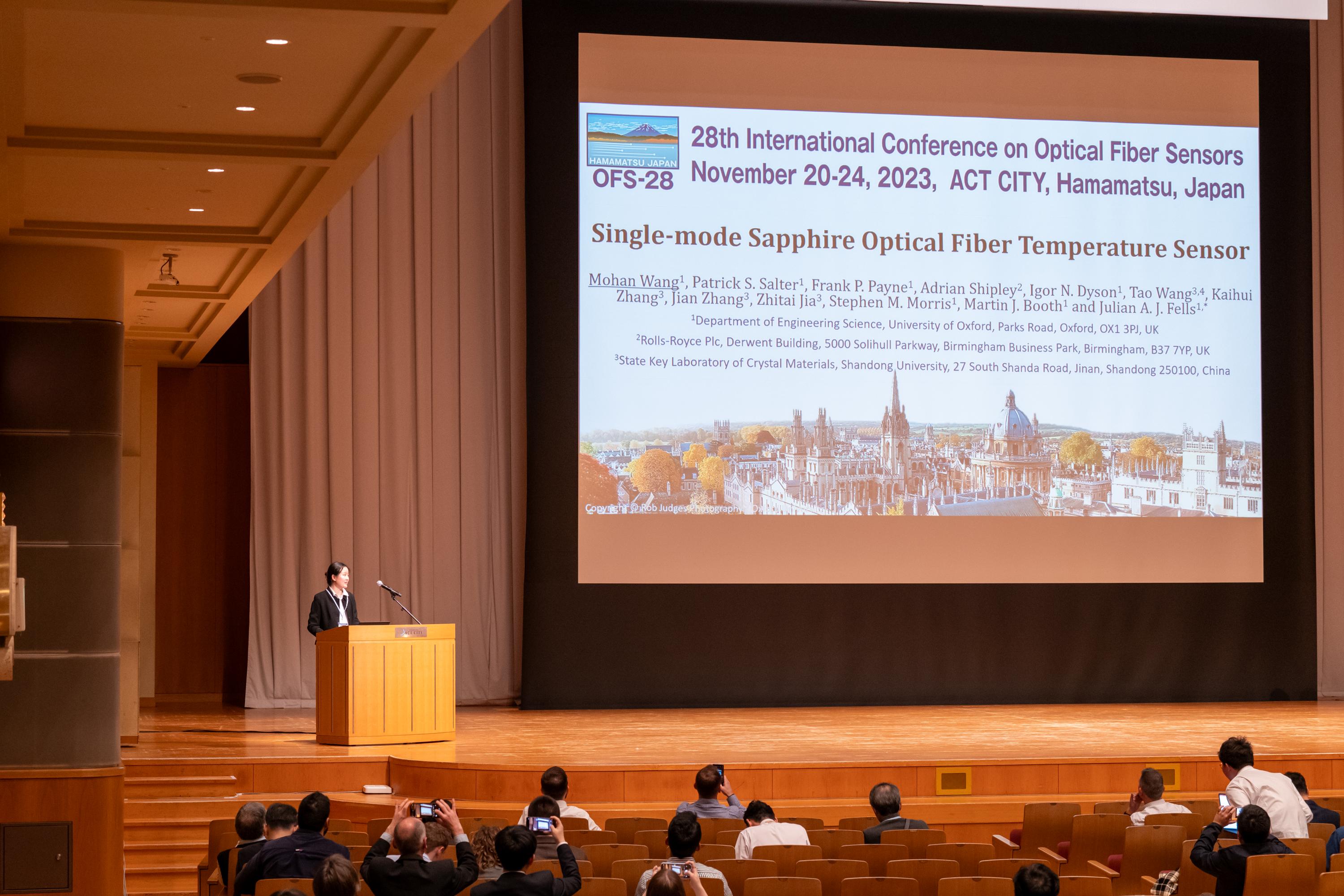  Dr Mohan Wang Presenting: an East Asian man is standing at a lectern of a large lecture room, presenting a PowerPoint which is shown on a large screen.