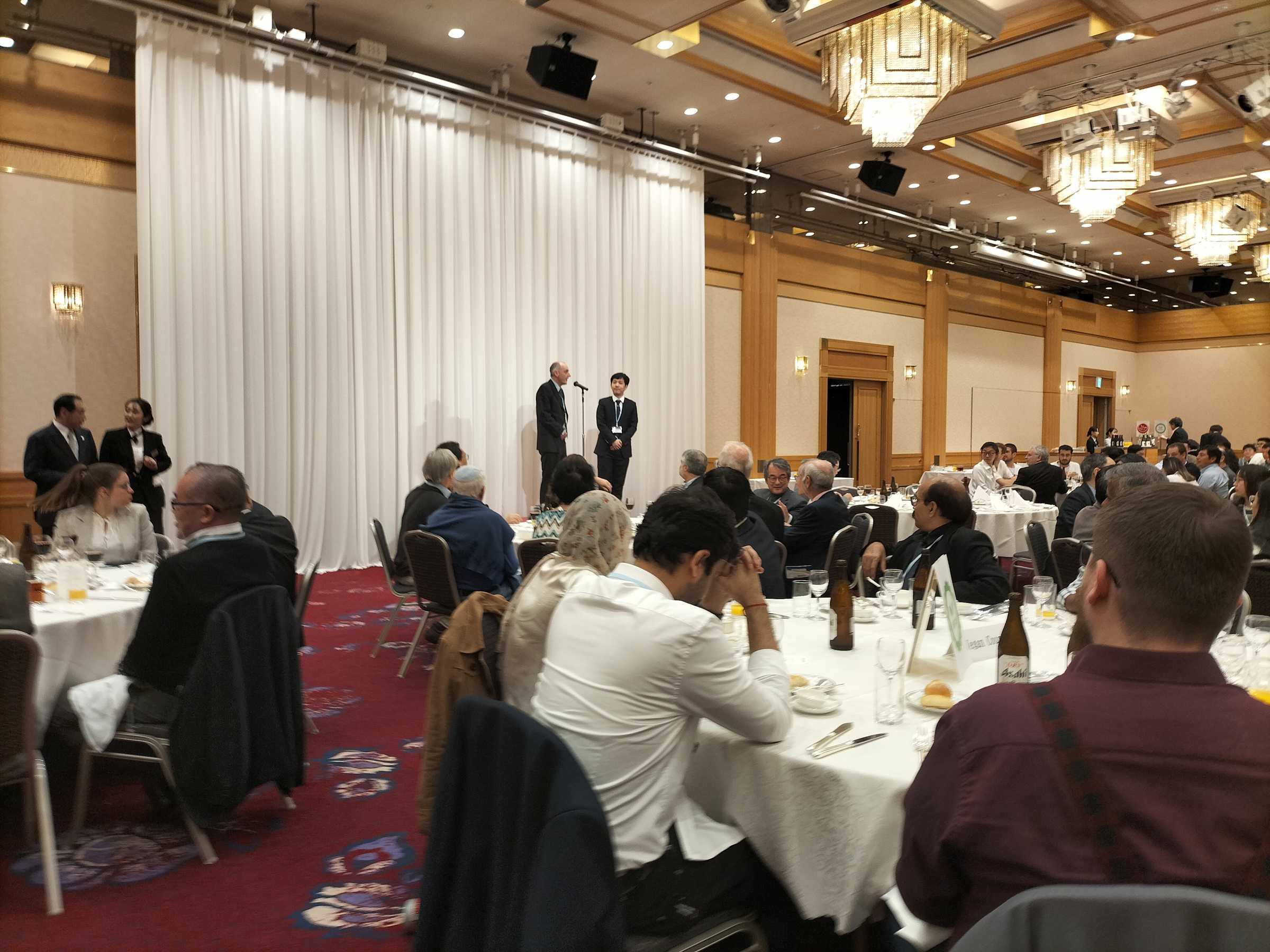  Acceptance Speech: an image of a white man at a mic on stage with an East Asian man. The stage is facing a room of people sat at dining tables.
